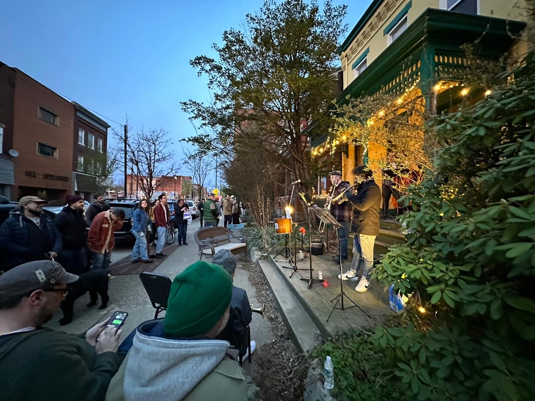 Image shows an audience gathered on the sidewalk in the early evening dancing and listening to a Dixieland jazz band performing on a green and yellow porch lit by white string lights
