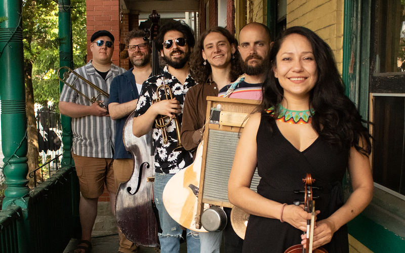 Six musicians gather on a yellow and green front porch, holding a trombone, double bass, cornet, guitar, washboard, and violin
