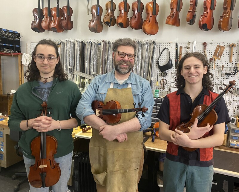 Three smilling luthiers holding violins stand in front of a workbench with an assortment of bows, violins, and tools displayed on a pegboard above