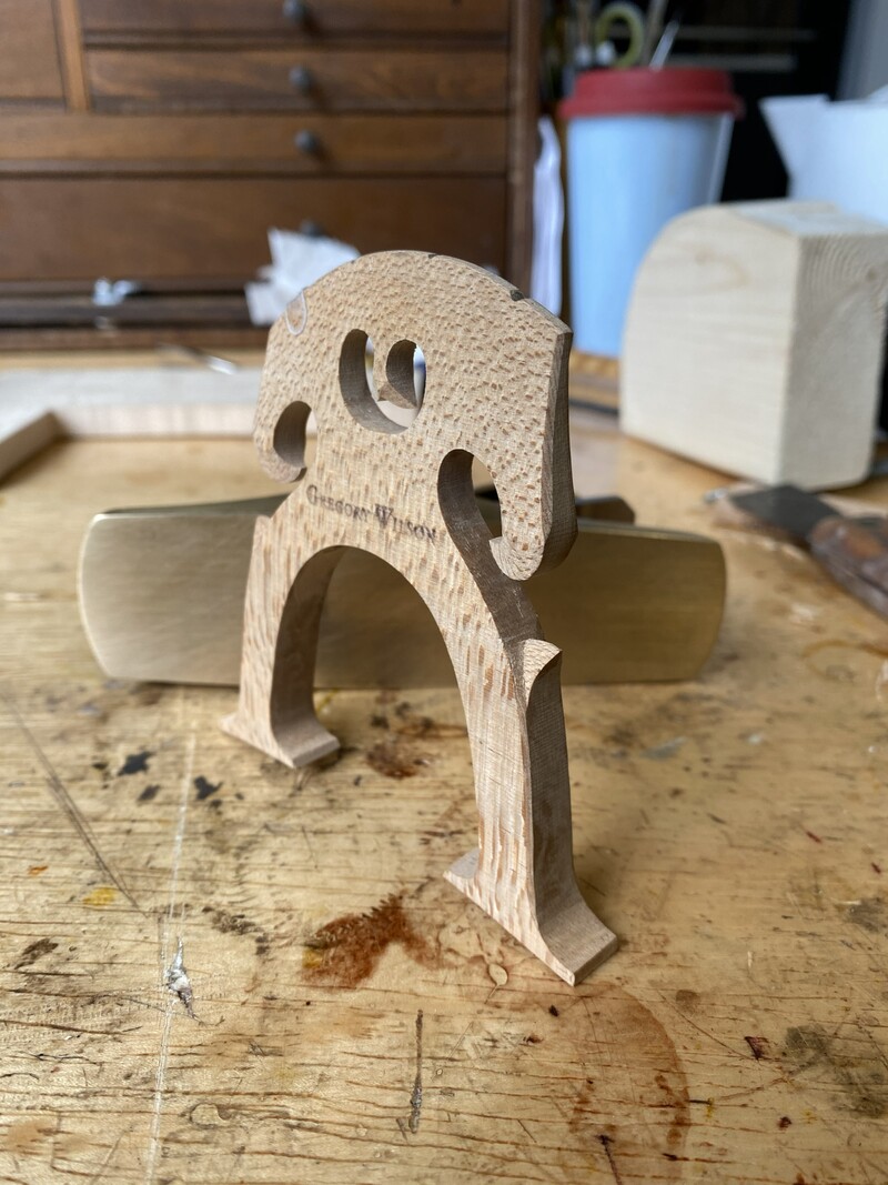 A Belgian cello bridge stands upright against a brass block plane on a worn workbench
