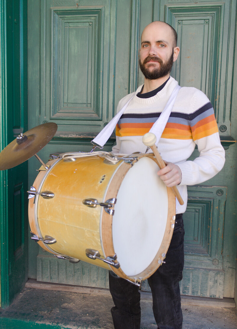 A smiling bearded musician in a rainbow striped sweater holds a marching bass drum in front of a green double door