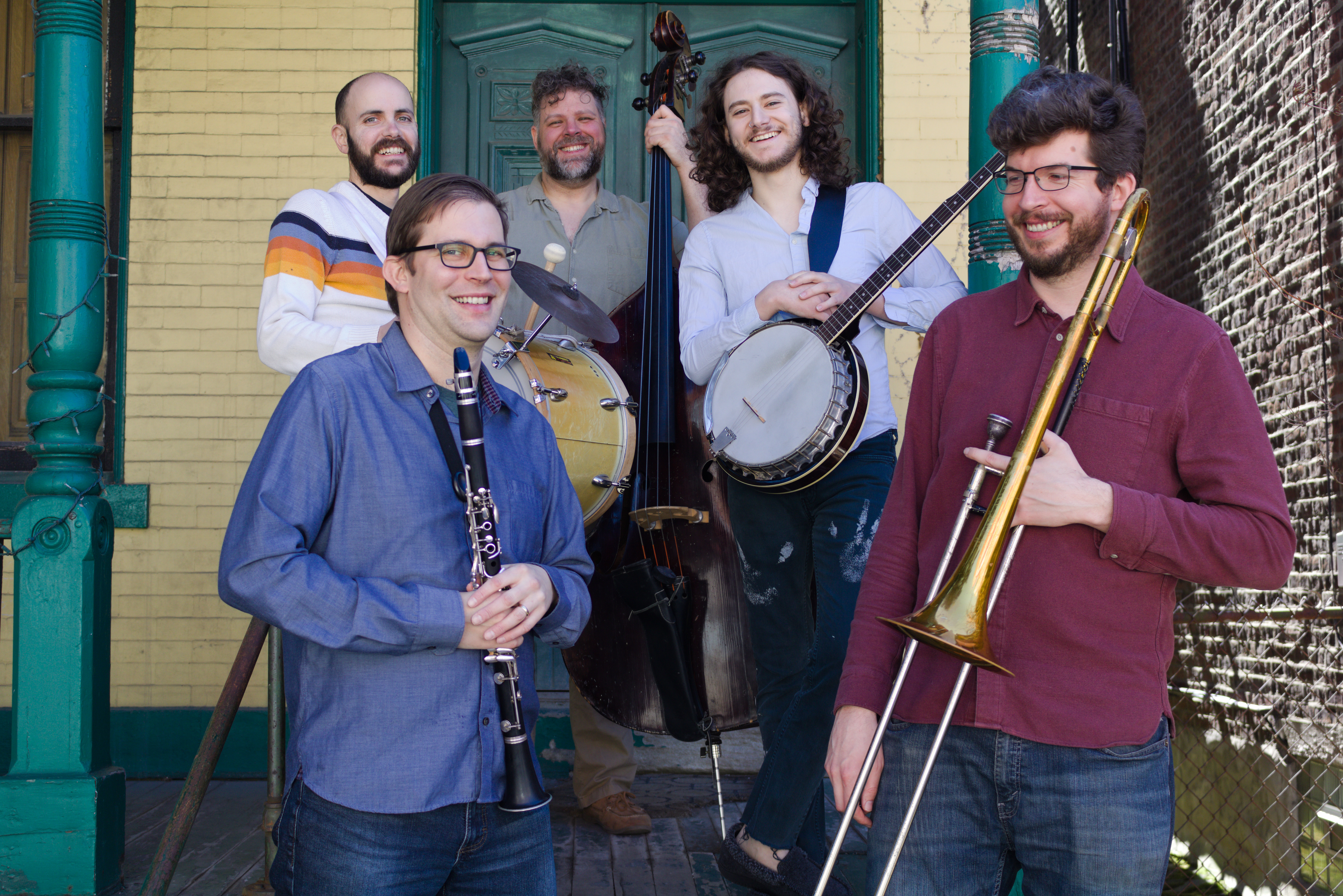 Image shows five smiling musicians gathered on the steps of a green and yellow porch holding a marching bass drum, a double-bass, a banjo, a clarinet, and a trombone