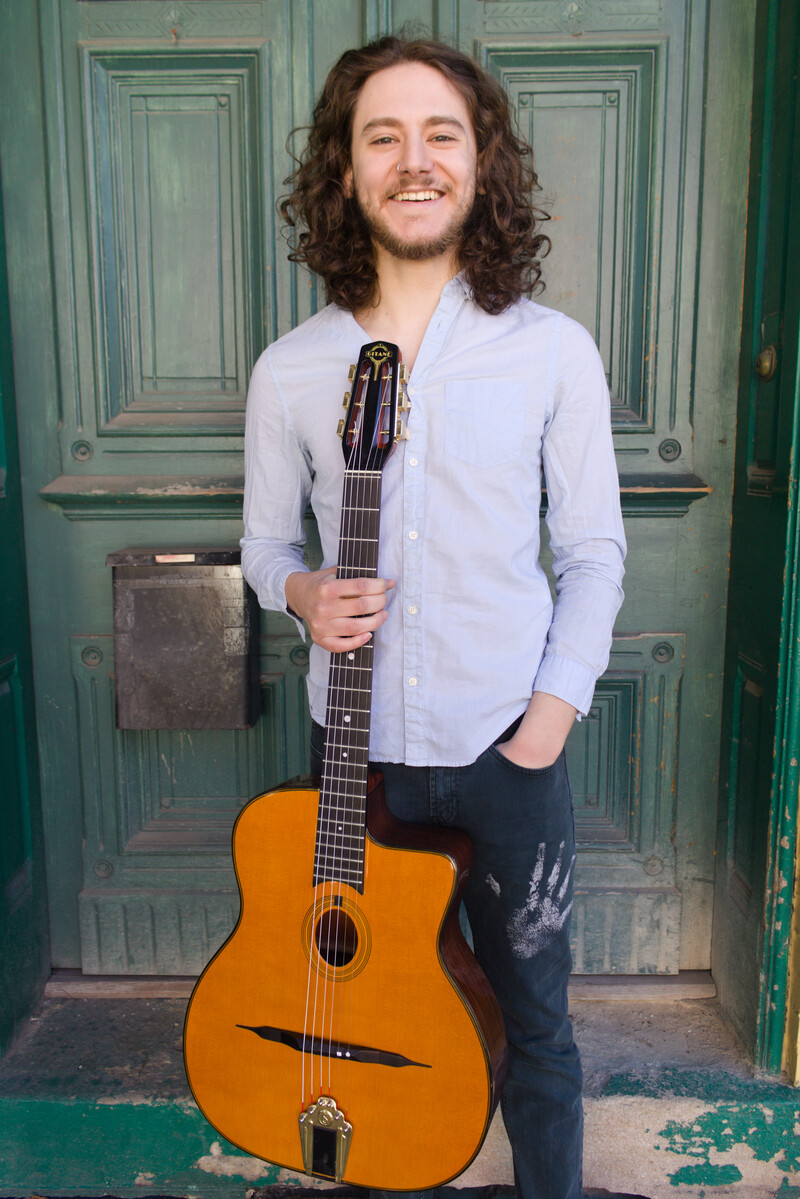 A smiling handsome young man with long curly brown hair holds a Selmer-Maccaferri  petite bouche guitar in front of a green double door