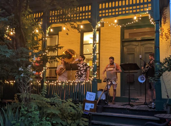 Five musicians perform on a yellow and green front porch with white string lights during the Unblurred art gallery crawl on Penn Avenue in Pittsburgh's Bloomfield Garfield cultural district