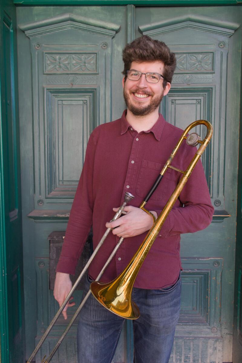 A smiling musician with brown hair and a beard stands in front of a green double door holding his trombone
