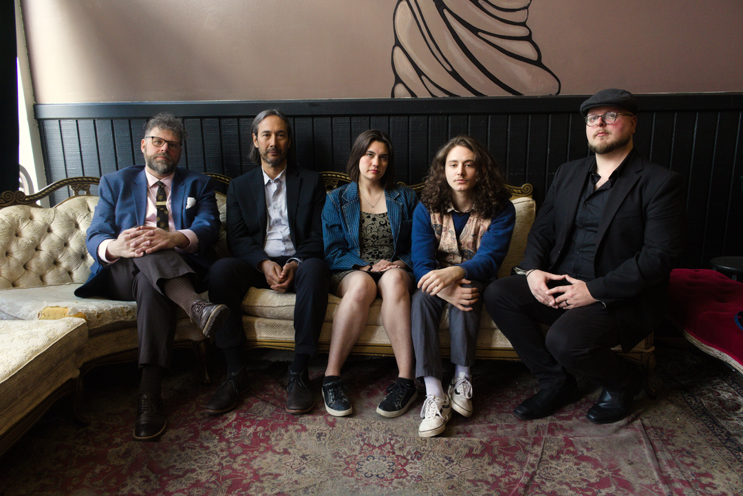Image shows five well-dressed musicians with serious expressions seated together on a vintage Victorian style white sofa