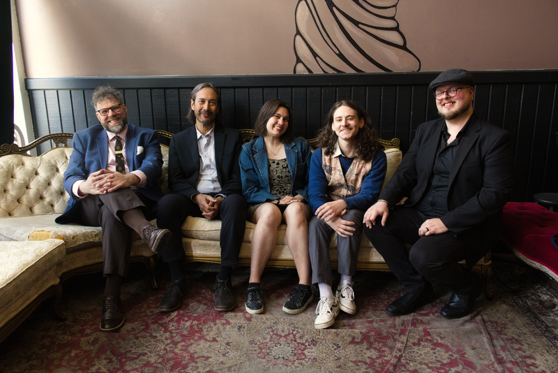 Image shows five musicians seated smiling on a sofa